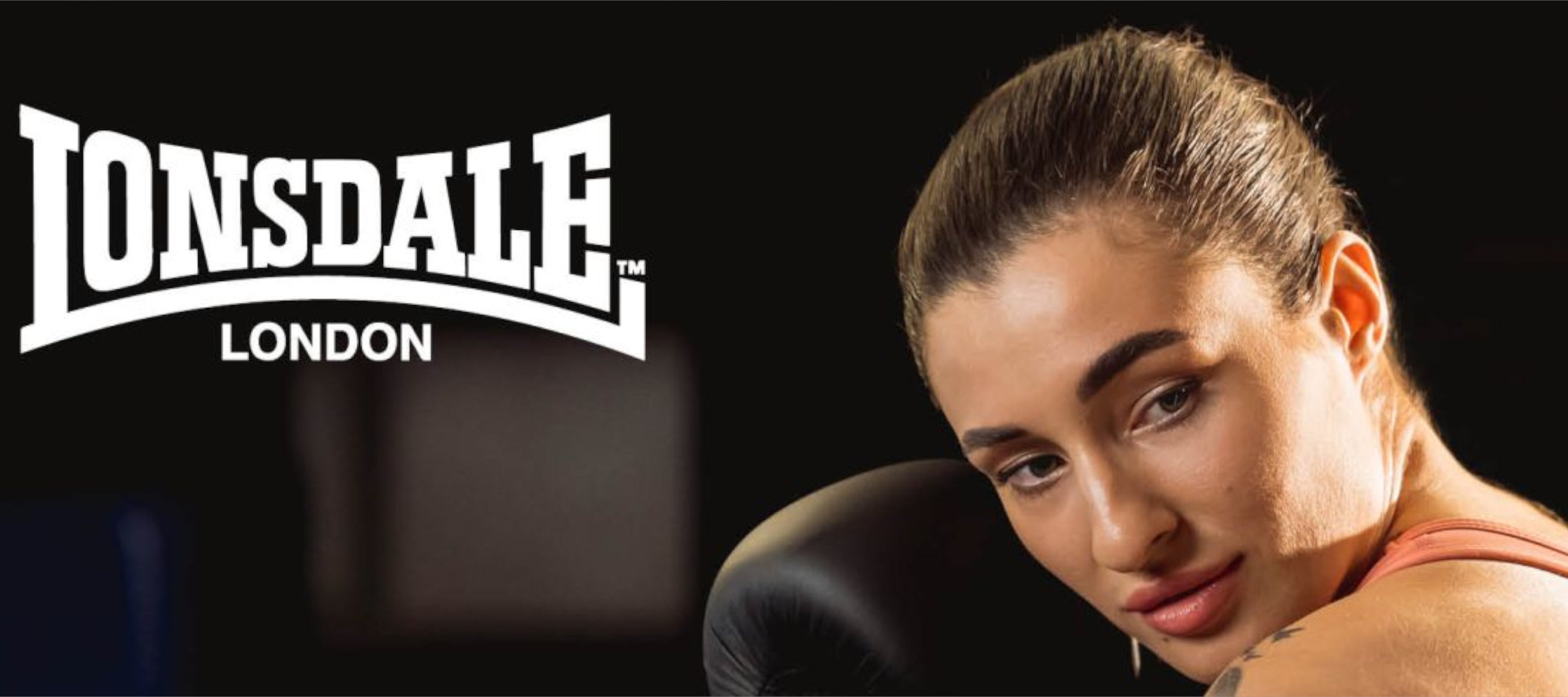 Frau mit Boxhandschuhen hält Deckung. Lonsdale Logo im Hintergrund.